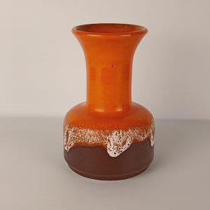 Può includere: Un vaso in ceramica con bordo svasato e collo cilindrico. La parte superiore è di un arancione vivace, che sfuma in un motivo a goccia di smalto bianco testurizzato che si sovrappone a una base solida di colore marrone scuro, creando contrasto.