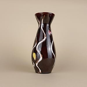 Peut inclure: Un vase en céramique brun foncé brillant avec un bord évasé et un col effilé. La surface présente des motifs verticaux en forme de gouttes blanches ondulées et une petite marque jaune, créant un contraste net avec le vernis brun profond.