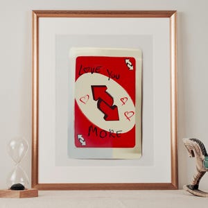 May include: Framed art print showing a red card with the words "Love You More" and a double arrow design. The card has a white oval with the text and is surrounded by hearts. The print is in a gold frame.