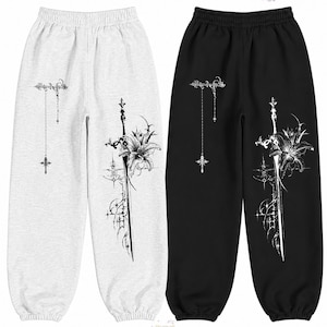 Op de afbeelding: Een lichtgrijze en een zwarte joggingbroek, beide met een elastische tailleband en boorden. De broekspijpen zijn voorzien van een witte grafische print van een zwaard verstrengeld met een leliebloem en een hangend kruisornament.