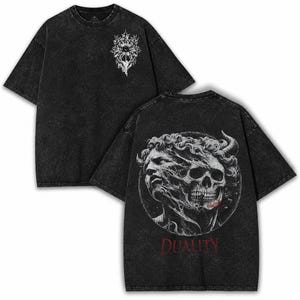 Puede incluir: Una camiseta negra con efecto lavado al ácido que presenta un pequeño gráfico floral blanco en el pecho. La parte posterior muestra un gran estampado circular de un rostro dividido, mitad humano y mitad calavera, con la palabra DUALITY en rojo.