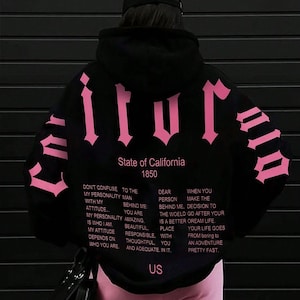 Puede incluir: Una sudadera negra con capucha que presenta grandes letras de estilo g&oacute;tico en color rosa en la espalda y las mangas. El texto dice State of California 1850, seguido de un p&aacute;rrafo de texto inspirador en varias columnas y las letras US en el dobladillo.