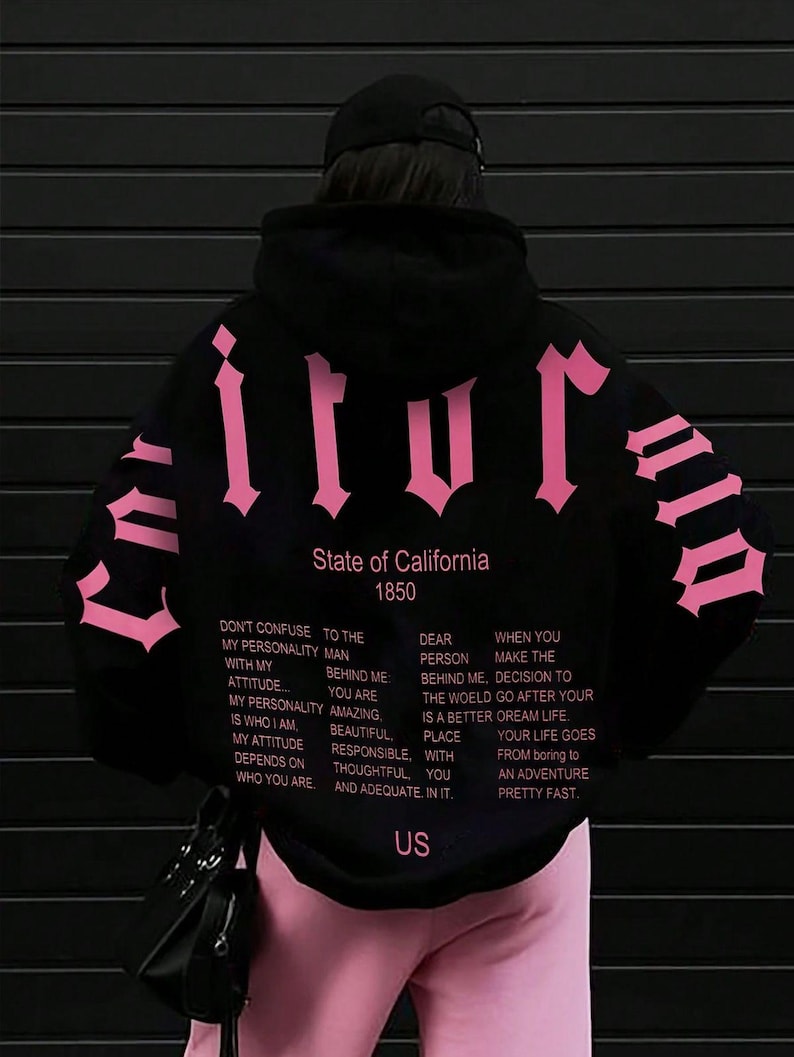 Puede incluir: Una sudadera negra con capucha que presenta grandes letras de estilo g&oacute;tico en color rosa en la espalda y las mangas. El texto dice State of California 1850, seguido de un p&aacute;rrafo de texto inspirador en varias columnas y las letras US en el dobladillo.