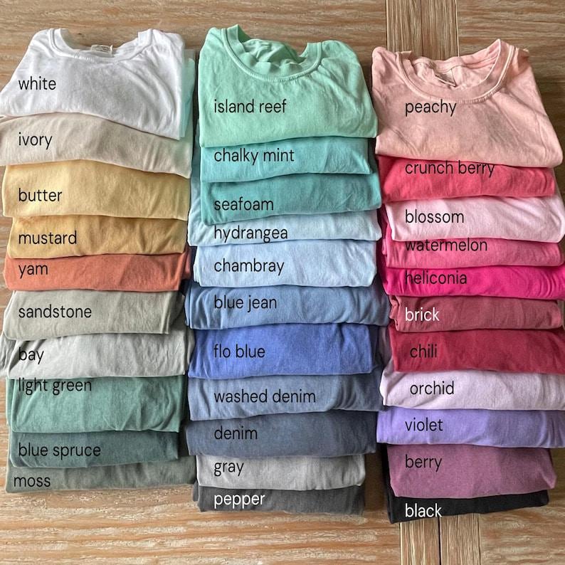 May include: A collection of folded crew neck t-shirts arranged in three vertical columns, featuring a wide variety of solid colours labelled with text including white, ivory, butter, mustard, yam, sandstone, bay, light green, blue spruce, moss, island reef, chalky mint, seafoam, hydrangea, chambray, blue jean, flo blue, washed denim, denim, grey, pepper, peachy, crunch berry, blossom, watermelon, heliconia, brick, chili, orchid, violet, berry, and black.