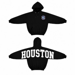 May include: A black hooded sweatshirt featuring a front kangaroo pocket and long sleeves. The front chest displays a circular NASA-style emblem, while the back features the word HOUSTON printed in large, bold, white capital block lettering.