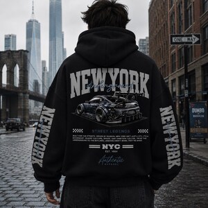 May include: A black hooded sweatshirt featuring a large graphic on the back with the text NEW YORK, a stylized sports car, and the phrase STREET LEGENDS. The sleeves also display the text NEW YORK in a vertical orientation in a light gray font.