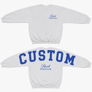 May include: A light grey long-sleeve sweatshirt featuring a blue Rush Soccer Club logo on the front chest. The back of the sweatshirt displays the word CUSTOM in large blue block letters above the blue Rush Soccer Club logo text.