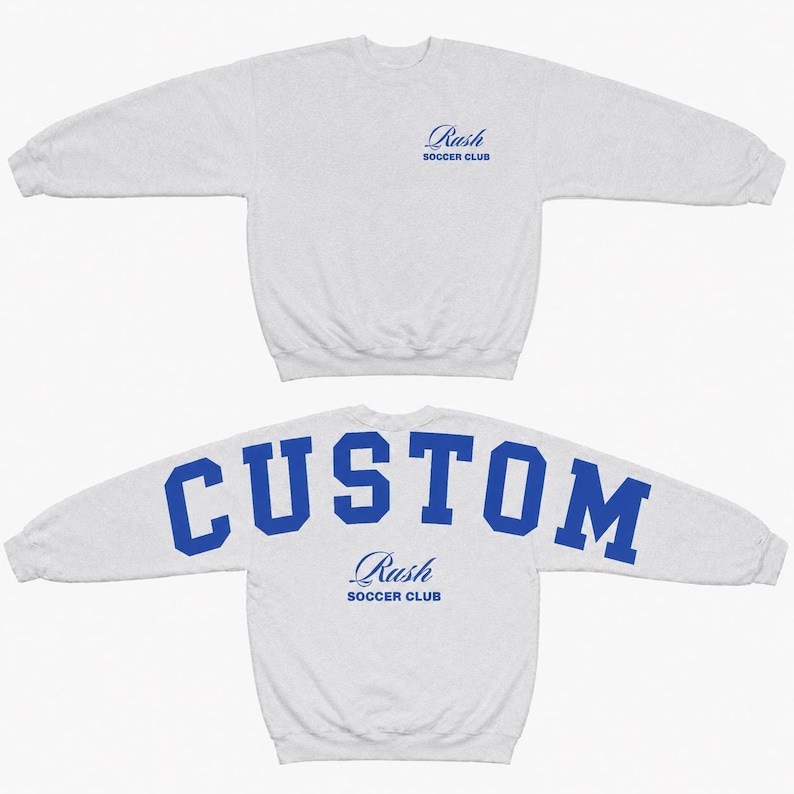 May include: A light grey long-sleeve sweatshirt featuring a blue Rush Soccer Club logo on the front chest. The back of the sweatshirt displays the word CUSTOM in large blue block letters above the blue Rush Soccer Club logo text.