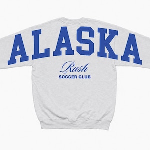Puede incluir: Una sudadera gris claro de manga larga con la palabra ALASKA en letras may&uacute;sculas azules grandes y negritas en la parte superior de la espalda. Debajo del texto, la palabra Rush est&aacute; impresa en fuente cursiva azul, seguida de SOCCER CLUB.