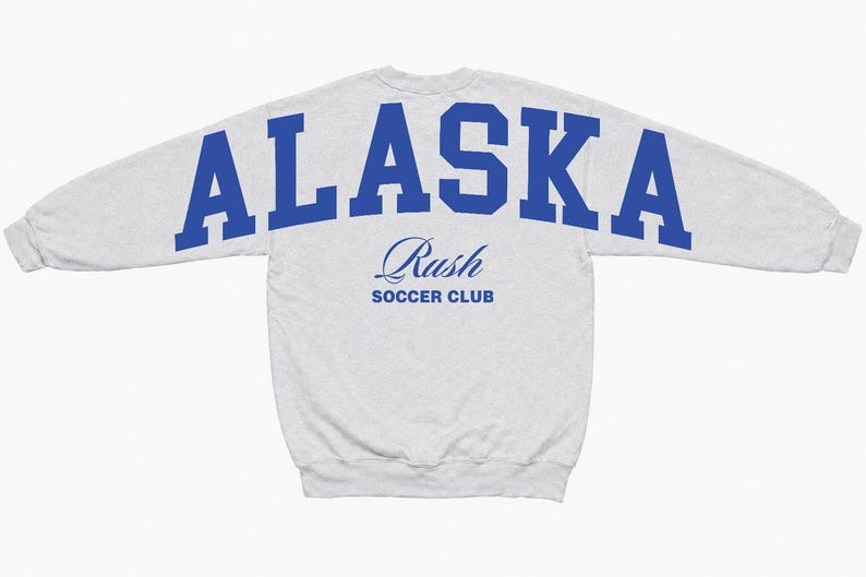 Puede incluir: Una sudadera gris claro de manga larga con la palabra ALASKA en letras may&uacute;sculas azules grandes y negritas en la parte superior de la espalda. Debajo del texto, la palabra Rush est&aacute; impresa en fuente cursiva azul, seguida de SOCCER CLUB.