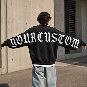 May include: A black long-sleeve sweatshirt featuring the white text YOURCUSTOM printed in a large gothic font across the back and extending onto the sleeves. The garment is worn over a white undershirt and light blue denim jeans.