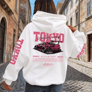 May include: A white hooded sweatshirt featuring a large graphic on the back with the text TOKYO Underground in pink, a stylized pink sports car illustration, Japanese characters, and the word Authentic Apparel in a script font.