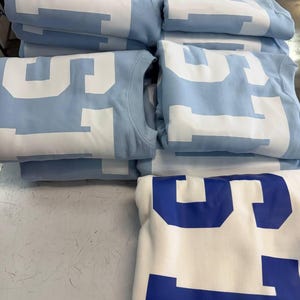 May include: A stack of folded light blue sweatshirts featuring large white block lettering that reads "STATE" across the front, alongside one folded white sweatshirt with large blue block lettering that also reads "STATE" on the front.