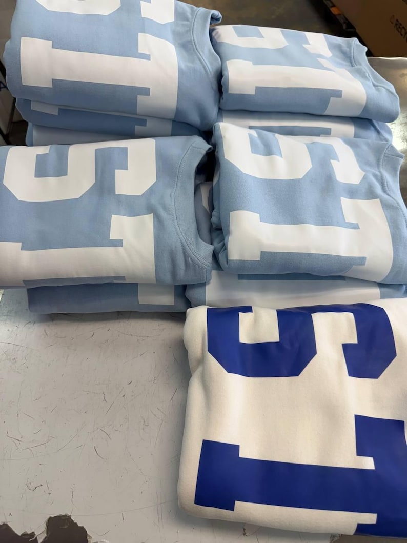 May include: A stack of folded light blue sweatshirts featuring large white block lettering that reads "STATE" across the front, alongside one folded white sweatshirt with large blue block lettering that also reads "STATE" on the front.