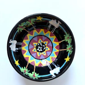 May include: A black ceramic bowl with a colorful, geometric design in the center. The bowl features white camels, palm trees, and yellow stars around the rim. The interior design includes red, yellow, blue, and purple shapes.