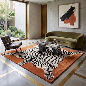 May include: A large rectangular area rug featuring a central black and white zebra stripe pattern on a solid burnt orange background. The rug is positioned on a smooth gray floor with gold inlay strips, situated beneath a glass coffee table.