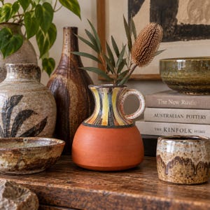 May include: A ceramic pitcher with a terracotta base and a neck featuring vertical stripes in yellow, black, and white. The pitcher has a single handle and contains a dried brown flower and green leaves, surrounded by other ceramic vessels and books.
