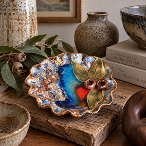 May include: A round, scalloped-edge ceramic dish featuring a vibrant glaze of deep blue, bright red, and earthy brown tones. The surface is decorated with three sculpted green leaves and two small brown seed pods attached to the centre.