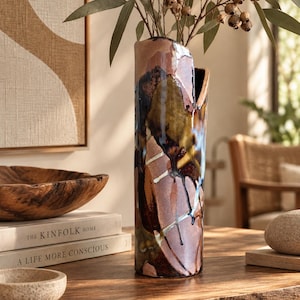 May include: A tall, cylindrical ceramic vase with a glossy, multi-coloured glaze featuring shades of brown, tan, deep blue, and white. The surface has an abstract, dripped paint effect with a distinct vertical cutout at the top rim.