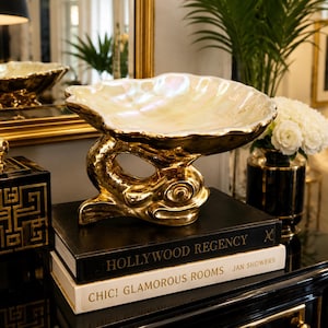 May include: A decorative bowl featuring a shell-shaped top with an iridescent finish, supported by a gold-coloured base sculpted in the form of a fish. The bowl rests on two stacked books titled Hollywood Regency and Chic! Glamorous Rooms.