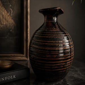 May include: A dark brown ceramic vase with horizontal textured lines. The vase has a wide, rounded body and a narrow, flared neck. The vase is on a dark marble surface.
