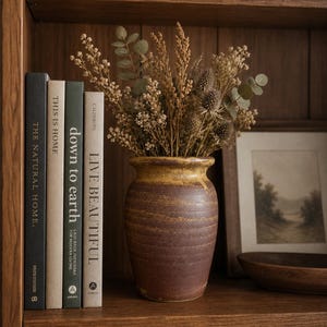May include: A rustic brown and yellow ceramic vase holds dried flowers and foliage on a wooden shelf. Books with titles including "This is Home" and "Live Beautiful" are placed beside the vase. A framed landscape print is also visible.