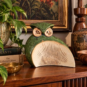 May include: A ceramic owl figurine featuring a speckled dark green glaze on the upper body and head, with a cream-coloured, semi-circular chest area decorated with fine, radiating dark brown etched lines and two round, light-coloured eyes.