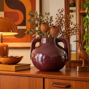 May include: A glossy, deep plum-coloured ceramic vase with two prominent side handles, filled with a bouquet of dried brown foliage and seed pods, positioned centrally on a wooden surface.