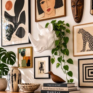 May include: A white ceramic rhinoceros head wall planter containing a trailing green pothos plant, positioned on a wall surrounded by various framed art prints, a wooden bird figurine, a ceramic vase, a bowl, and a stack of three books.
