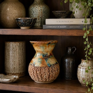 May include: A collection of ceramic pottery displayed on wooden shelves, including a central vase with a textured, rough-hewn base and a smooth, glazed neck in shades of amber, blue, and cream, surrounded by various stoneware vessels and books.