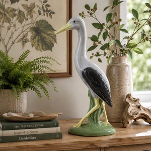 May include: A ceramic heron figurine featuring a light grey neck and head, a long yellow beak, black wings, and yellow legs. The bird stands on a green base, captured in a glossy finish with detailed sculpting of its feathers and posture.