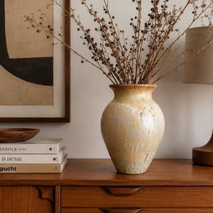 May include: A ceramic vase with a rounded body and a flared rim, featuring a glossy glaze that transitions from a warm golden-yellow at the top to a muted, speckled cream and grey tone at the base, containing a cluster of dried brown branches.