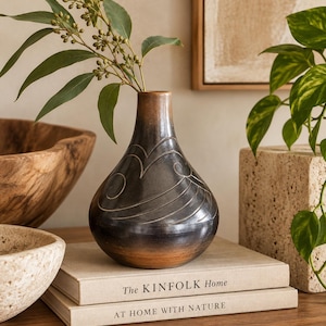 May include: A dark brown, pear-shaped ceramic vase with a smooth, glossy finish and subtle carved wave patterns. The vase features a narrow neck and a wider base, with a gradient of lighter brown tones near the bottom, holding a green eucalyptus branch.