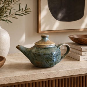 May include: A ceramic teapot with a mottled blue and green glaze and a contrasting tan lid. The teapot features a curved handle, a tapered spout, and a rounded body, showcasing a textured, handcrafted appearance with visible colour variations.