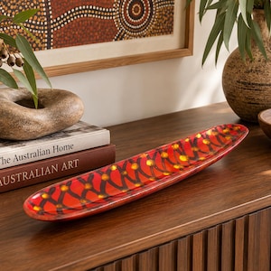 May include: An elongated, oval-shaped decorative tray featuring a vibrant red base colour. The surface is adorned with a repeating pattern of dark brown, intersecting lines and small, bright yellow circular dots throughout the design.
