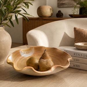 May include: A shallow, wide ceramic bowl with a wavy, scalloped rim and a glossy, marbled beige and cream finish. The bowl contains three brown pears. The piece features a smooth, curved design and sits on a flat surface next to stacked books.