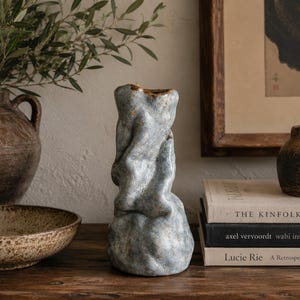 May include: A tall, hand-formed ceramic vase with an irregular, undulating surface texture and a mottled light blue and grey glaze. The vessel features a narrow opening at the top and a wider, bulbous base, showcasing a unique, organic sculptural shape.