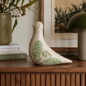 May include: A cream-coloured ceramic bird figurine featuring a distinct crackle glaze finish and embossed green fern leaf patterns on its side. The bird is depicted in a seated position with a smooth, rounded head and a pointed tail.