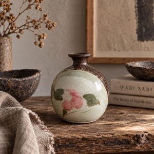 May include: A small, round ceramic vase with a narrow neck, featuring a crackled light green glaze and a hand-painted pink and green floral motif. The upper portion and base of the vase are finished in a dark, speckled brown textured glaze.