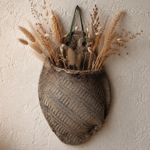May include: A wall-mounted ceramic pocket planter with a dark, mottled grey finish and a textured, diagonal ribbed pattern. The vessel is suspended by a thin green cord and contains an arrangement of dried beige grasses and wheat stalks.