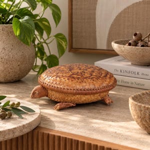 May include: A turtle-shaped lidded box crafted from woven light brown rattan and wicker. The shell features a darker brown speckled pattern, while the head and four small feet are detailed with coiled rattan. The box has a rounded, textured surface.