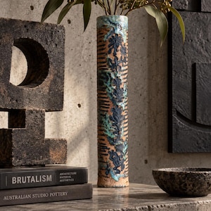May include: A tall, cylindrical ceramic vase with a textured, light brown clay body featuring horizontal incised lines and a vertical, raised decorative pattern in dark blue and turquoise glaze, holding a sprig of green eucalyptus leaves.