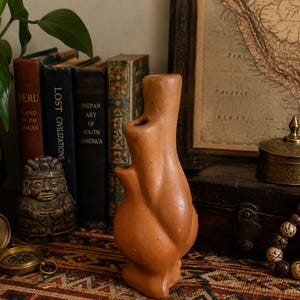 May include: A terracotta-coloured ceramic vessel with a dual-spout design, featuring a rounded base and a tall, slender neck. The surface is smooth and matte, showcasing a natural clay finish with subtle variations in tone and texture.