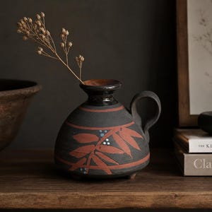 May include: A black ceramic vase with a handle, adorned with red leaf patterns and blue dots. The vase contains a dried plant stem. The vase is on a wooden surface, with a large bowl and books in the background.