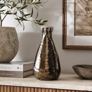 May include: A teardrop-shaped ceramic vase with a metallic bronze and dark brown glazed finish. The vase features a narrow neck with a textured, off-white rim. The surface has a reflective, uneven quality with visible vertical ridges.