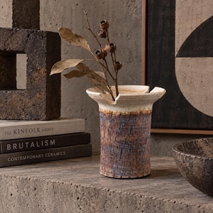 May include: A cylindrical ceramic vase with a flared, notched rim. The upper portion is off-white, while the lower body features a textured, bark-like finish in shades of brown, rust, and blue-grey, containing a dried branch with small seed pods.