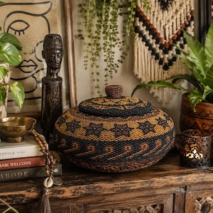 May include: A round, lidded basket featuring a dense, intricate pattern of black and golden-brown beads. The basket has a low-profile, wide shape with a small, beaded knob on the centre of the lid, showcasing detailed craftsmanship and texture.