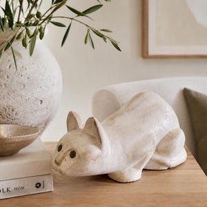May include: A cream-coloured, textured cat figurine in a crouching position with hollow circular eyes and a smooth, matte finish. The sculpture features a rounded back and pointed ears, crafted with a weathered, off-white appearance.