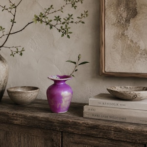May include: A small, vibrant purple vase with a flared rim, positioned between a speckled beige bowl and a stack of three books titled The Kinfolk Home, at 100, and Wabi-Sabi Welcome, topped with a shallow, textured beige bowl.