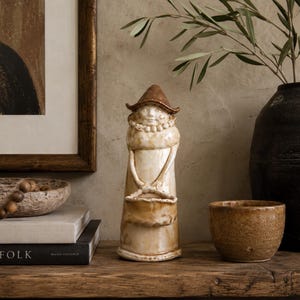 May include: A ceramic figurine of a person wearing a wide-brimmed brown hat and a ruffled collar, holding a basket in front of their body. The figure has a glossy, cream-coloured glaze with brown accents and a smooth, rounded, elongated form.